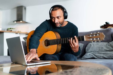 Siyah adam evde gitar çalıyor ve dizüstü bilgisayar kullanıyor.