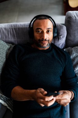 40 yaşında bir adam evdeki kanepede dinleniyor ve telefonuyla müzik dinliyor.