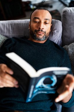 Karışık ırk adamı evdeki kanepede uzanıp kitap okuyor.