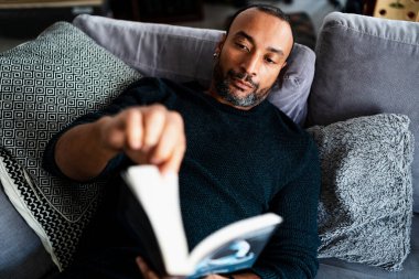 40 yaşında bir adam kanepede oturup kitap okuyor.