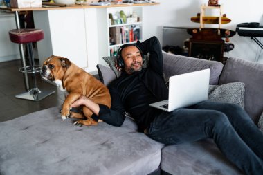 Orta yaşlı bir adam kanepede dinleniyor ve köpeğinin yanında müzik dinliyor.