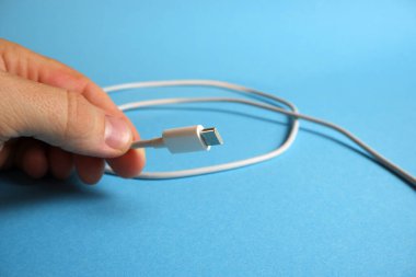 Parmaklar mavi arkaplanda C tipi arayüzlü beyaz bir usb kablosu tutuyor..