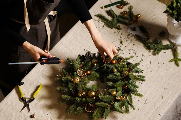 Yapıştırıcı tabancalı çiçekçi elleri keten masa örtüsünde Xmas ladin çelengi yapıyor. El yapımı eko dekorasyon. Dekor elementleri: Sarı altın cam toplar, koniler, yıldız anason, kurutulmuş kireç ve turuncu kupalar..