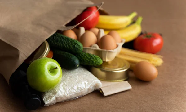 Makarna, pirinç, yağ, sirke, yumurta, muz, domates, reçel, salatalık, baharat. Tarafsız bej arka planda yiyecek malzemeleri olan kağıt torba. Karantina, bağış, koronavirüs konsepti için gıda teslimatı