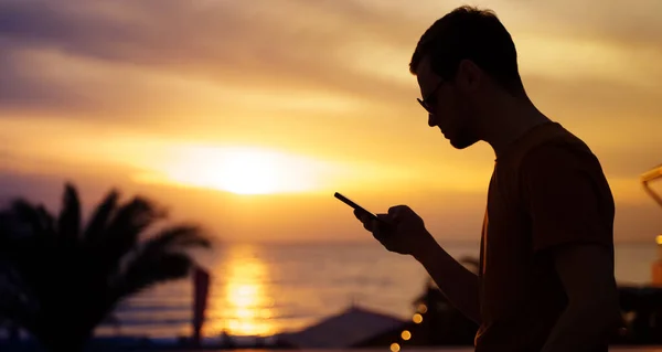 Profile of youn man standing at sea coast, browsing news or doing shopping using mobile phone. Banner, copy space