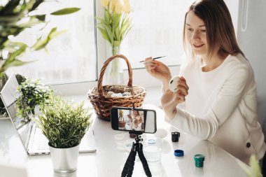 Genç kadın blogcu çevrimiçi canlı yayın yapıyor. Paskalya yumurtalarının nasıl süsleneceğini gösteriyorum. Akıllı telefonlu tripod, yumurtalı sepet, boya ve dizüstü bilgisayar. COVID tecrit kavramı sırasında geleneksel tatiller