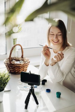 Genç kadın blogcu çevrimiçi canlı yayın yapıyor. Paskalya yumurtalarının nasıl süsleneceğini gösteriyorum. Akıllı telefonlu tripod, yumurtalı sepet, boya ve dizüstü bilgisayar. COVID tecrit kavramı sırasında geleneksel tatiller