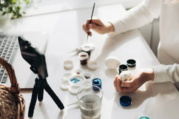 Genç kadın blog yazarının elleri canlı yayında. Paskalya yumurtalarının nasıl süsleneceğini gösteriyorum. Akıllı telefonlu tripod, yumurtalı sepet, boya ve dizüstü bilgisayar. COVID tecrit kavramı sırasında geleneksel tatiller