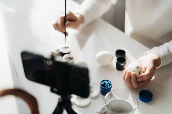 Genç kadın blogcu çevrimiçi canlı yayın yapıyor. Paskalya yumurtalarının nasıl süsleneceğini gösteriyorum. Akıllı telefonlu tripod, yumurtalı sepet, boya ve dizüstü bilgisayar. COVID tecrit kavramı sırasında geleneksel tatiller