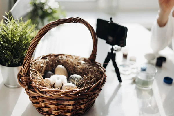 Blog yazarı, Paskalya yumurtası dekorasyonu üzerine canlı yayın yapıyor. Akıllı telefonlu tripod, yumurtalı sepet, boya tüpleri. COVID tecrit kavramı sırasında geleneksel tatiller