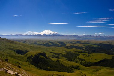 Platodan sönmüş volkana ve Elbrus Dağı 'nın karlı zirvesine mükemmel bir manzara. Aşağıda yeşil bir vadi var.
