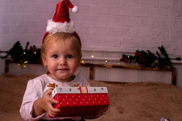 Una ragazza in un abito di Capodanno apre un regalo, la ragazza non guarda la fotocamera seduta sul letto con le palle di Natale e la decorazione di Natale — Foto Stock