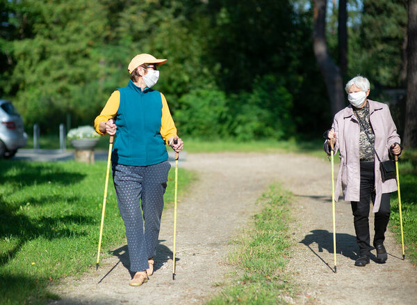 Two senior women wearing medical masks walking with nordic walking poles during covid-19 pandemic