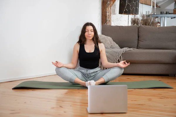 Yoga online. Relaxed young woman meditating front of laptop at home, sitting in lotus position and doing breathing exercises