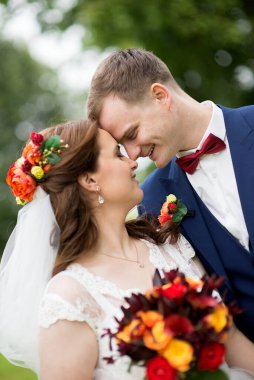 Düğün çifti yapraklı yeşil çalıların arasında dururlar, gelin elinde renkli bir buket tutarken, her ikisi de, doğal ışık ve duyguyu yakalayan samimi, huzurlu bir portre çizerler..