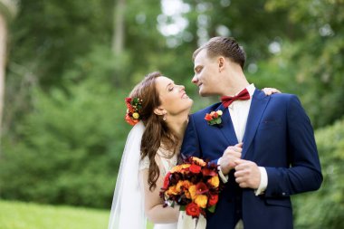 Düğün çifti yapraklı yeşil çalıların arasında dururlar, gelin elinde renkli bir buket tutarken, her ikisi de, doğal ışık ve duyguyu yakalayan samimi, huzurlu bir portre çizerler..