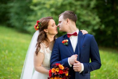 Düğün çifti yapraklı yeşil çalıların arasında dururlar, gelin elinde renkli bir buket tutarken, her ikisi de, doğal ışık ve duyguyu yakalayan samimi, huzurlu bir portre çizerler..