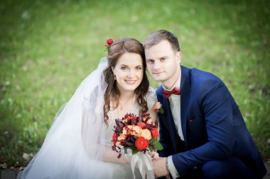 Düğün çifti yapraklı yeşil çalıların arasında dururlar, gelin elinde renkli bir buket tutarken, her ikisi de, doğal ışık ve duyguyu yakalayan samimi, huzurlu bir portre çizerler..