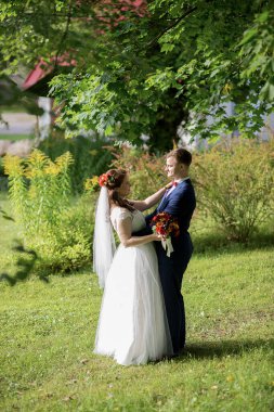 Düğün çifti yapraklı yeşil çalıların arasında dururlar, gelin elinde renkli bir buket tutarken, her ikisi de, doğal ışık ve duyguyu yakalayan samimi, huzurlu bir portre çizerler..
