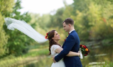 Gelin duvağı rüzgarda süzülürken, yeni evli çift sakin suyun yanında sarılıp hareketli, sıcak ve karşılıklı güven dolu bir düğün anı yaratıyor..