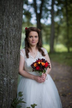 Genç gelin elinde renkli bir buketle bir ağacın yanında duruyor. Yumuşaklık, denge ve yansıtıcı ruh haliyle dolu sakin ve zarif bir düğün portresi oluşturuyor..