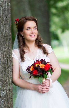 Genç gelin, bir ağacın yanında bir buket çiçek tutarken düşünceli bakışlarını kaçırarak doğal derinlik ve sessiz romantik duygularla huzurlu bir düğün portresi oluşturuyor..