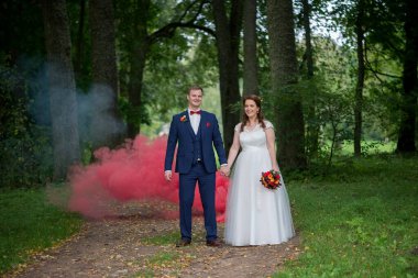 Gülümseyen gelin ve damat birlikte yeşil orman yolunda yürürken arkalarında kırmızı duman kayboluyor, neşeyi ve kutlamayı simgeliyor..