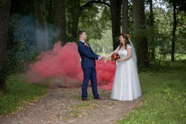 Damat gelinin eline uzanırken, kırmızı duman orman yolunu arkalarından dolduruyor.