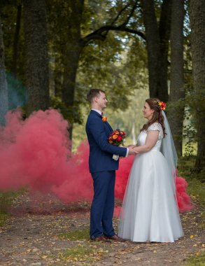 Gülümseyen gelin ve damat, kırmızı duman arkalarına yayılıp neşeli bir düğün atmosferi yaratırken bir orman yolunda el ele tutuşuyorlar..