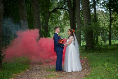 Gelin ve damat gölgeli bir orman yolunda sakince dikilirken kırmızı duman arkalarında sürükleniyor, doğadaki tutkuyu ve kutlamayı simgeliyor..