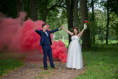 Genç düğün çifti kollarını zaferle kaldırırken kırmızı duman arkalarında dalgalanırken zafer, mutluluk ve açık hava düğün günlerinde paylaştıkları enerjiyi ifade ediyorlar..