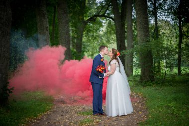 Genç gelin ve damat ormanda nazik bir öpücük paylaşırken kırmızı duman arkalarında sürükleniyor ve romantik ve dramatik bir açık hava düğün anı yaratıyor..