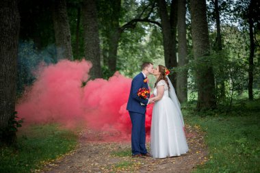 Genç evliler, arkalarından yayılan kırmızı dumanla, kutlama, tutku ve doğal yeşilliği birbirine karıştırarak, yumuşak bir şekilde öpüşürler..