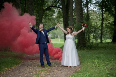 Genç gelin ve damat ellerini kaldırarak ve parlak gülümsemelerle kutluyor arkalarında kırmızı duman kaybolurken, neşeyi, birliği ve güçlü ortak bir düğün anısını simgeliyor..