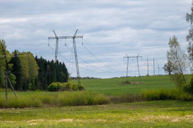 Yeşil Letonya çayırları boyunca uzanan yüksek voltaj kuleleri orman kenarları ve bulutlu bahar gökyüzü ile çevrilidir..