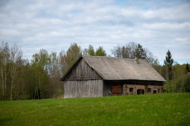 An aged wooden barn with stone lower walls stands on a vivid green Latvian meadow, framed by mixed spring trees beneath a textured cloudy sky.