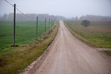 Düz çakıl yolu açık tarım tarlalarından geçiyor. Elektrik direkleri ve uzak ağaçlar Letonya 'nın sessiz kırsallarında buğunun içinde kayboluyor..