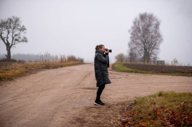 Orta yaşlı bir kadın, Letonya 'nın kırsal kesimlerinde sis, tarlalar ve çıplak ağaçlarla çevrili sonbahar kırsalını fotoğraflayan sessiz bir toprak yolda duruyor..