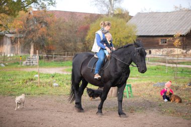 Çiftlik binaları ve evcil hayvanların yanında ata binen bir anne ve bebeğin yatay görüntüsü. Ilık sonbahar ışığı aile değerlerini taşra geleneklerini ve açık havada huzurlu yaşamı taşır..