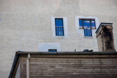 Eski ahşap bir çatının üzerinde mavi yansımalı küçük üst kat pencereleri zıt malzemelerle katmanlı bir şehir mimari sahnesi oluşturur..