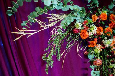 Horizontal image of vibrant flower arrangement with roses greenery and branches placed on rich burgundy fabric background.