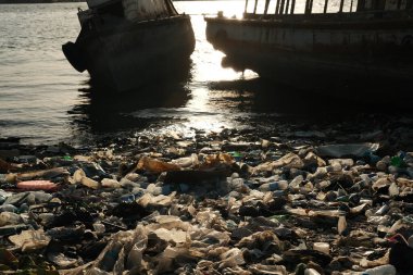 Kirlenmiş bir kıyı şeridi, atılmış plastik atıklar ve döküntüler, arka planda iki terk edilmiş tekne, su yüzeyine yansıyan güneş ışığı.