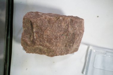 Kırmızımsı kahverengi tonu olan pürüzsüz, cilalanmamış bir kaya, beyaz bir yüzey üzerinde dinleniyor, doğal dokusu ve mineral bileşimini sergiliyor.