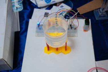 Etrafında kablolar ve elektronik bileşenler olan sarı bir tabana yerleştirilmiş, suyla dolu şeffaf bir konteynır, bir bilim projesi veya deneyi gösteriyor.