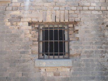 Parmaklıklı pencereli yıpranmış taş bir duvar, kırsal dokular ve tarihsel mimari sergiliyor.