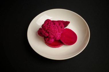 Canlı pembe karnabahar ve siyah arka planda beyaz bir tabakta yuvarlak pembe badem kurabiyelerinin yer aldığı yaratıcı bir tatlı sunumu.