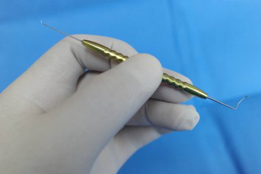 Altın saplı ve kıvrımlı metal uçlu bir diş aletini tutan bir el, mavi arka planda.