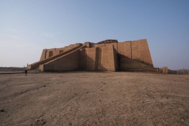 Çorak bir arazide antik ziggurat yapısı, açık mavi gökyüzünün altında basamaklı mimari ve çamur tuğlalı yapı sergileniyor.