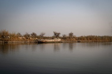 Yıpranmış, geleneksel bir ahşap balıkçı teknesi Shatt el-Arap Su Yolu 'nun çamurlu kıyı şeridinde demirli ve dinleniyor..