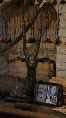 Bronz bir antilop heykeli, tarihi bir figürün çerçeveli fotoğrafı ve arka planda asılı renkli püsküllerin yer aldığı dekoratif bir sergi.